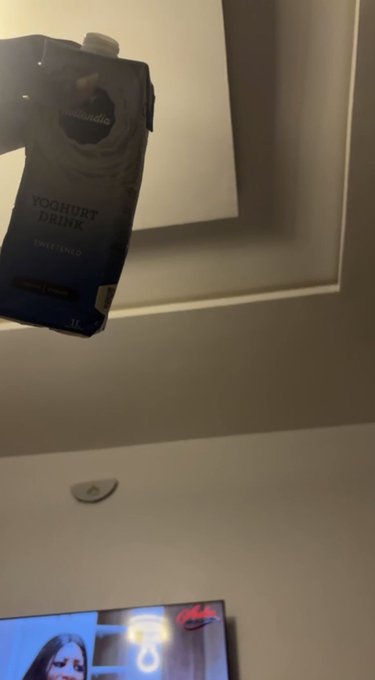 A hand holds up a blue 'essential Waitrose YOGHURT DRINK' carton in front of a recessed wall niche, with a TV visible at the bottom of the frame.