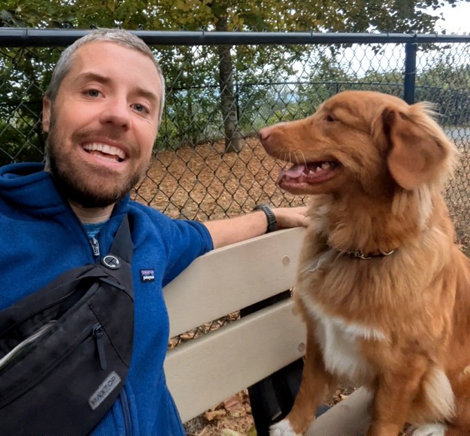 A smiling bearded man in a blue Patagonia jacket and Maxpedition sling bag sits on a park bench beside a happy golden-furred dog, with a chain-link fence in the background.