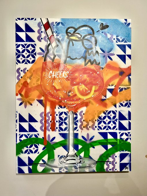 A mixed-media artwork featuring a wine glass with an orange cocktail that says 'C-HEERS,' set against a blue and white geometric tile background with two bird figures, a straw, and an orange slice.