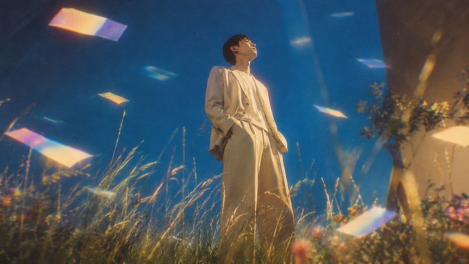 A person in a tan trench coat stands in a moonlit field with grass and flowers, gazing upward as colorful rectangular light streaks float around them like ethereal portals, evoking the mystical and dreamy aesthetic of the Num collection mint announcement. The surreal scene complements the launch message, symbolizing the magical unveiling of partner passes and upcoming rarity utility reveals.