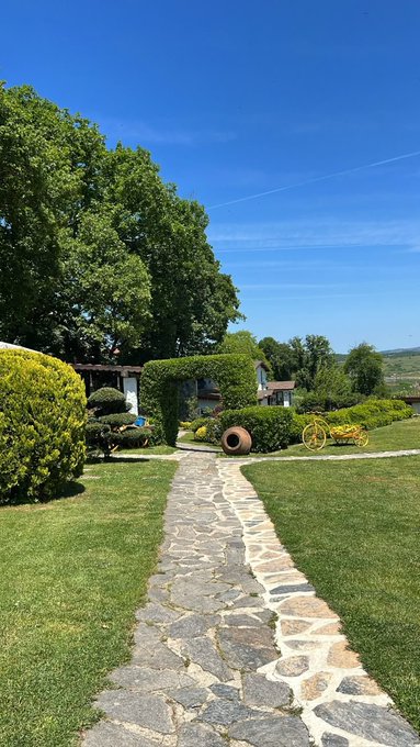 A stone pathway winds through a lush green garden with manicured hedges and topiaries, leading toward a building under a clear blue sky.