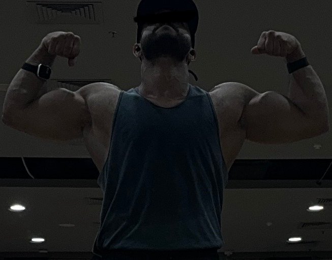 A muscular man wearing a cap and dark tank top flexes his biceps in a double bicep pose, photographed from a low angle that emphasizes his muscular build, while the accompanying post playfully compliments his impressive physique gained from gym workouts.