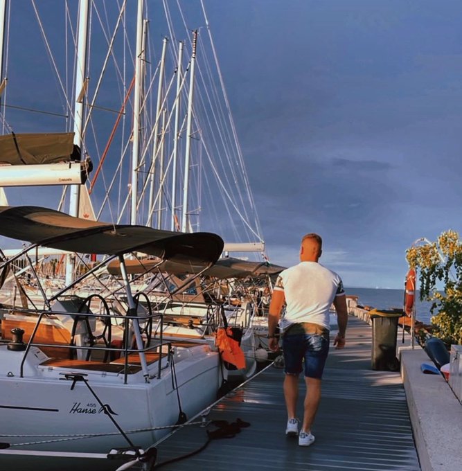 The image shows a man walking along a pier, carrying an orange bag. There are several boats docked at the pier, including a sailboat with its sails down. The background features a cloudy sky, adding to the overall atmosphere of the scene. The caption "Grand rising solana enjoyers" suggests that this location might be popular for sailing or boating activities in the area.