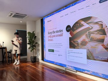 A man stands in a room with a large screen displaying a webpage with the text "Keep the stories only you would know" and a photo of people in traditional dress.