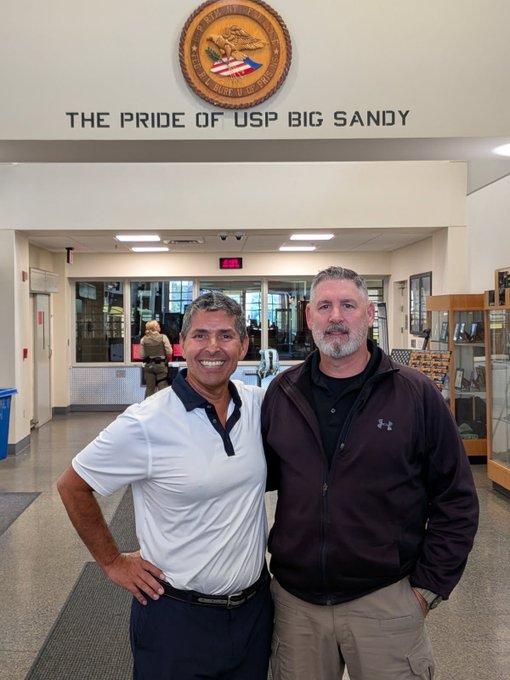 Two men pose smiling in an institutional lobby beneath a sign reading 'THE PRIDE OF USP BIG SANDY' with a United States Penitentiary seal.