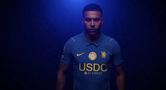 A promotional image of a male soccer player wearing a blue Chelsea FC jersey with the USDC by Circle sponsor logo on the front, illuminated by blue lighting.