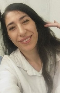 A selfie of a smiling woman with long dark hair wearing a white shirt, posing with a peace sign.