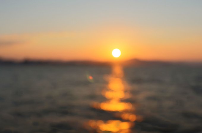A blurry photograph captures a sunset over the ocean, with the sun's golden light creating a shimmering reflection on the water's surface.