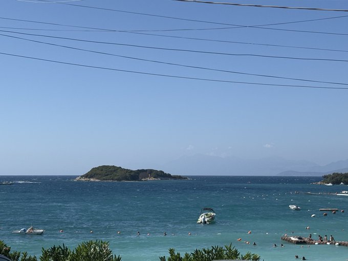 A small, lush tropical island sits in clear turquoise waters with boats anchored nearby and a mountainous coastline in the distance, suggesting a Mediterranean or Adriatic beach destination—a fitting backdrop for the poster's boast about enjoying an extended vacation while the crypto market surges.