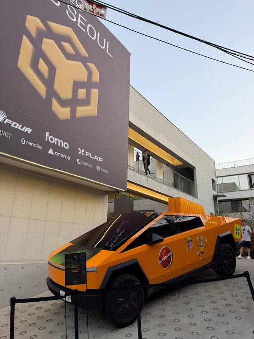 An orange, futuristic-looking vehicle is parked outdoors in front of a large building with a billboard displaying "SEOUL" and logos including "FOUR," "fomo," and "FLAP".