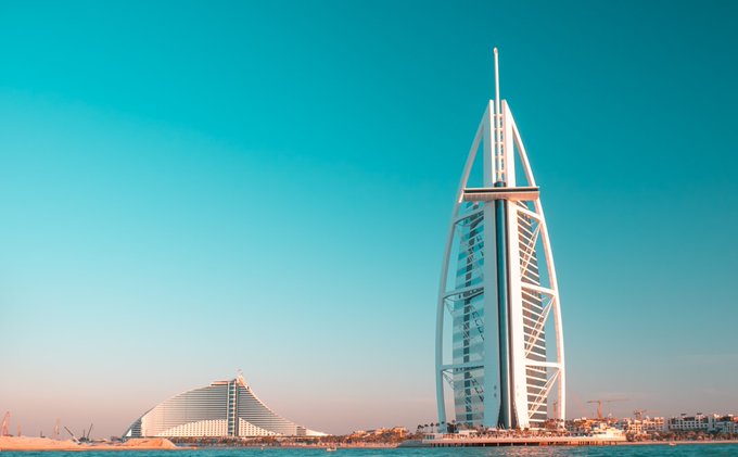 The Burj Al Arab hotel stands prominently against a clear blue sky, with the Jumeirah Beach Hotel visible in the background.