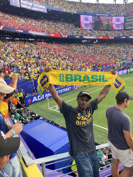 A fan at MetLife Stadium holds up a bright yellow Brazil scarf during the FIFA World Cup, with the stadium's massive crowd of 83,000 fans visible behind him and signage reading "New York New Jersey" overhead, perfectly matching the post's excitement about repping $DOG at the World Cup 2026.