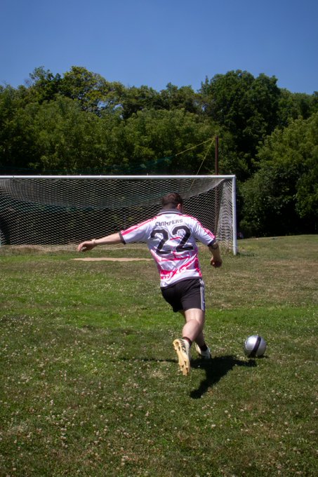 A man in a white and pink jersey with the number 22 and the name "Cuttrello" on the back runs toward a soccer ball on a grassy field, with a goal and trees in the background.