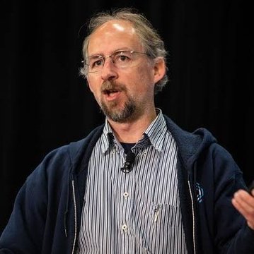 A man with glasses, a beard, and a striped shirt under a dark hoodie speaks at an event, gesturing with his hand. The image relates to the post featuring Adam Back, a prominent figure in the Bitcoin space, sharing his bullish prediction about Bitcoin reaching seven figures if the US establishes a Strategic Bitcoin Reserve.