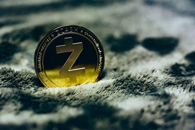 Close-up photograph of a physical gold and silver Zcash (ZEC) cryptocurrency coin standing upright on a textured fabric surface.