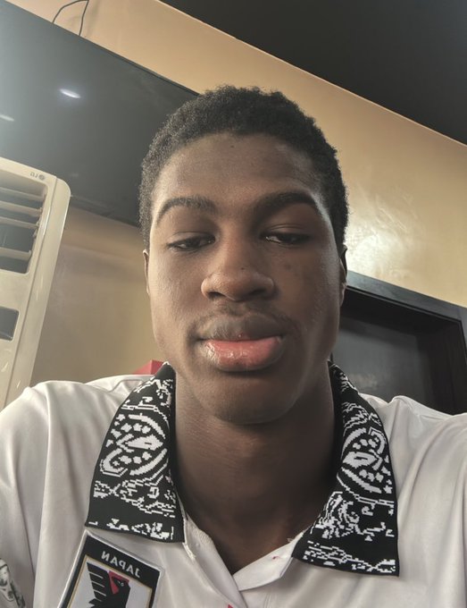 The image features a man wearing a white shirt with black and white patterns, including a bandana design. The man has short hair and is looking directly at the camera while making a funny face for the photo. The background of the image shows a TV screen mounted on the wall behind the man.