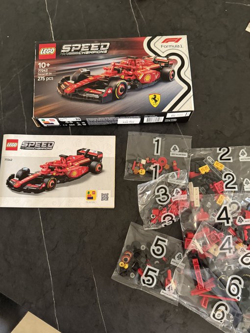 A LEGO Speed Champions Formula 1 set (set number 77243, 275 pcs) for ages 10+ is laid out on a dark countertop, showing the box, instructions, and six numbered bags of red and black pieces.