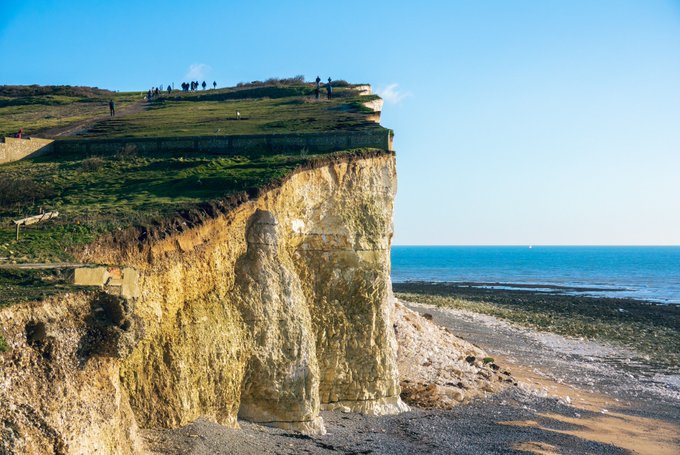 In this image, a cliff face with white rock formations can be seen overlooking the ocean. A group of people are standing at the edge of the cliff, enjoying the view and taking pictures. The sky above them appears blue, suggesting that they might be on a sunny day. The scene captures the beauty of nature and the excitement of the moment shared by these individuals as they admire the stunning landscape before them.