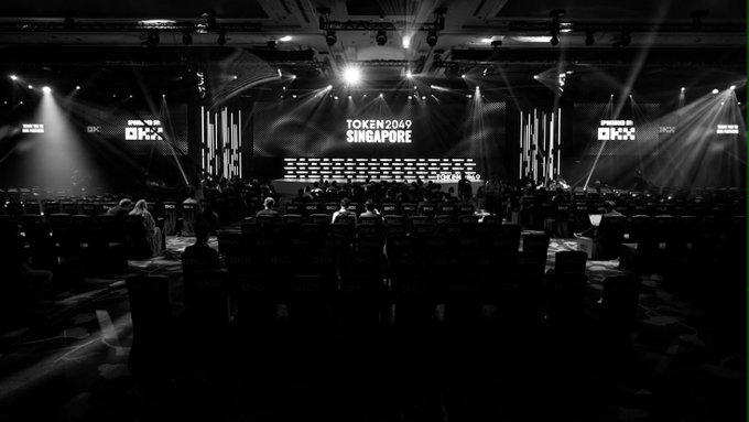 The black and white image shows a dimly lit conference hall before the TOKEN2049 Singapore event begins, with the stage displaying "TOKEN2049 SINGAPORE" and "OKX" branding, setting the scene for the networking and product showcase that will take place at OKX's Booth PB5-70.