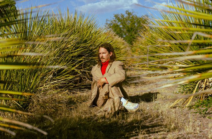 A person with long hair, wearing a beige coat and red turtleneck, crouches in dry grass and is framed by out-of-focus palm fronds in the foreground.