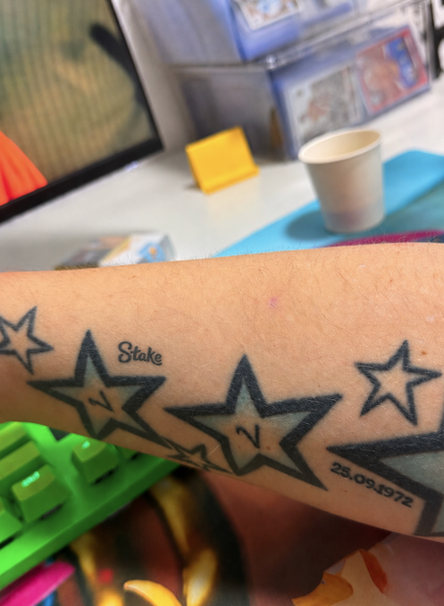A close-up of a forearm tattoo featuring black-outlined stars with the word 'Stake' and the date '25.09.1972'.