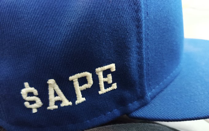 A close-up of a blue baseball cap featuring white embroidered text that reads '$APE'.