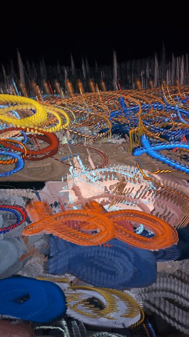 A chaotic, glitch-style digital artwork with colorful, distorted shapes resembling coiled snakes or tentacles in yellow, blue, and orange against a dark background.