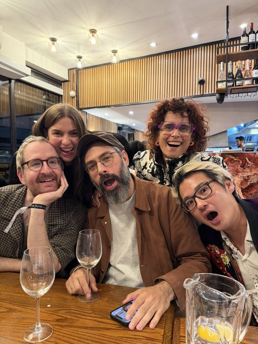 A group photo of five people smiling and posing enthusiastically around a wooden table with wine glasses and a pitcher of water in a restaurant setting.