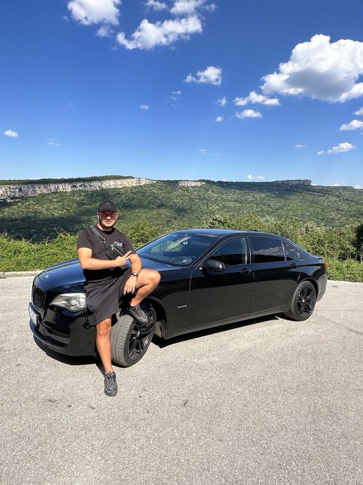 A man leaning against a black BMW sedan, posing for a photo at a scenic overlook with mountains and a blue sky in the background.