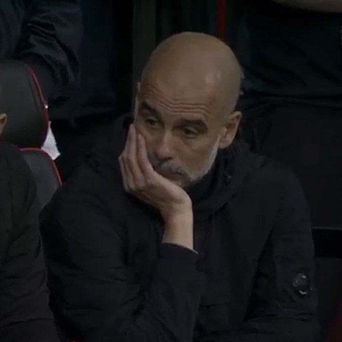 A bald man in a dark hoodie sitting on a bench with his hand resting on his cheek, looking thoughtful.