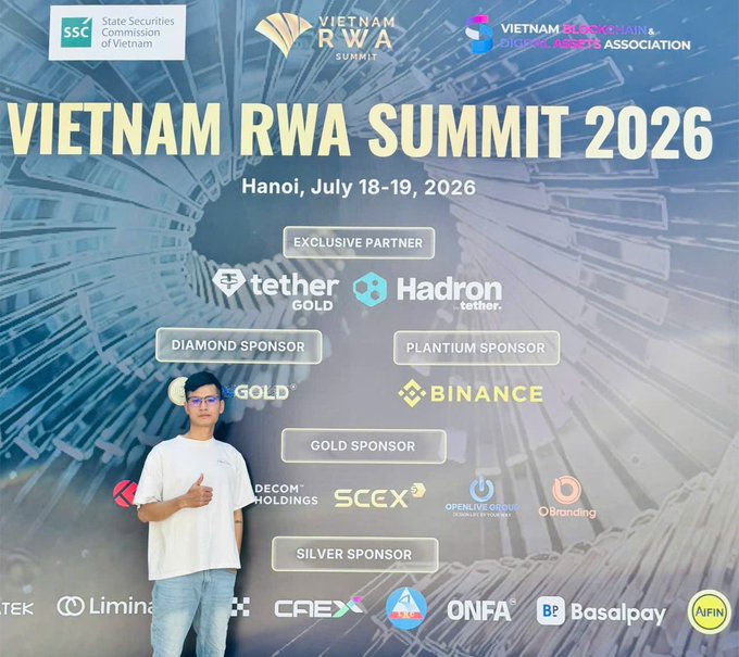 A promotional photo of a person posing in front of the 'Vietnam RWA Summit 2026' event banner (Hanoi, July 18-19, 2026), featuring sponsors including Tether Gold, Hadron, Binance, and Decorm Holdings.