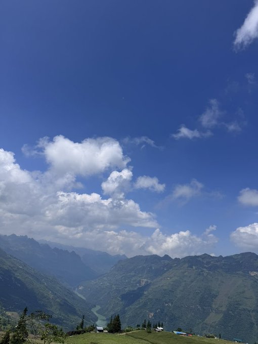 A scenic mountain landscape with green valleys, a winding river, and white clouds against a blue sky.