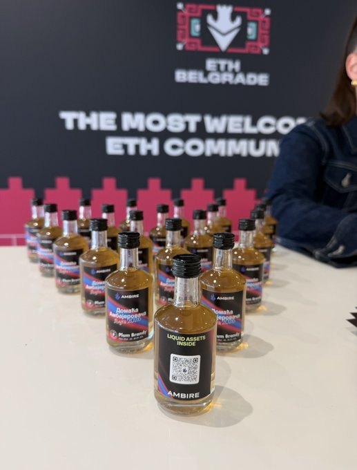 Multiple small bottles of an amber liquid labeled "AMBIRE" and "Banana Anejoed Style" with "LIQUID ASSETS INSIDE" and a QR code are arranged on a table at the "ETH BELGRADE" event with a banner reading "THE MOST WELCO(MING) ETH COMMUN(ITY)."
