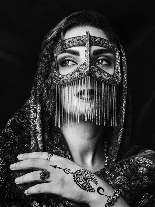 A black-and-white portrait of a woman wearing an ornate beaded veil/headpiece and henna-decorated hands, gazing directly at the camera with intricate jewelry visible.