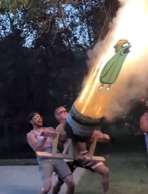 Two men in pink tank tops operate a wooden cannon that shoots a large fireball outdoors, humorously overlaid with a cartoon pickle character flying through the flames.