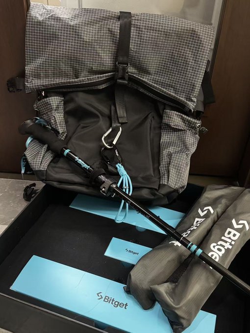 A Bitget-branded checkered hiking backpack with a matching trekking pole rests on a display tray featuring the "Bitget" logo, showcasing promotional gifts from the Bitget Mid-Autumn Festival giveaway. The accompanying post celebrates these outdoor-themed items as a creative way to encourage the crypto community to explore beyond their screens, with the branded accessories perfectly tying into the messaging.