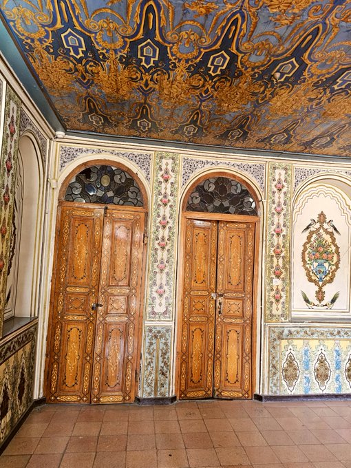 An ornate interior featuring intricately painted walls and a ceiling with floral and arabesque patterns, centered around two large, decorative wooden doors.