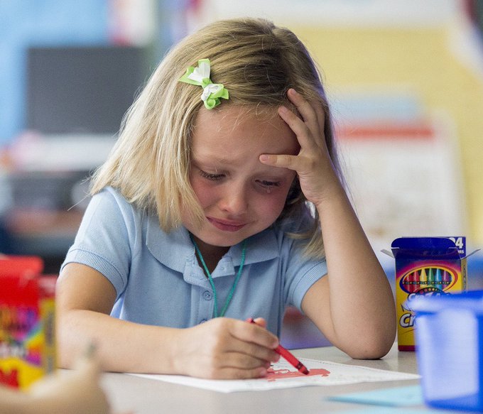 A young blonde girl with a green ribbon in her hair, wearing a light blue polo shirt, concentrating while drawing with a red colored pencil at a desk.