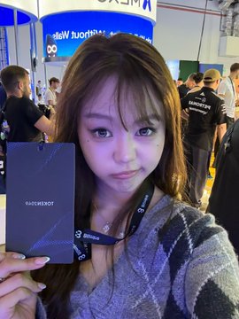 A young woman with long brown hair poses for a selfie at the GMGM main venue, holding a promotional card while wearing a lanyard, surrounded by event attendees.