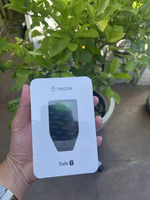 A hand holding a white box containing a Trezor Safe 7 hardware cryptocurrency wallet, displayed in front of a leafy green potted plant.