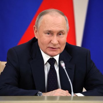 A man in a dark suit and tie sits at a desk with a microphone, positioned in front of Russian flags, suggesting a formal address or meeting. The accompanying post indicates this relates to Russia's push to expand cryptocurrency access, with the figure potentially being President Vladimir Putin promoting this initiative.