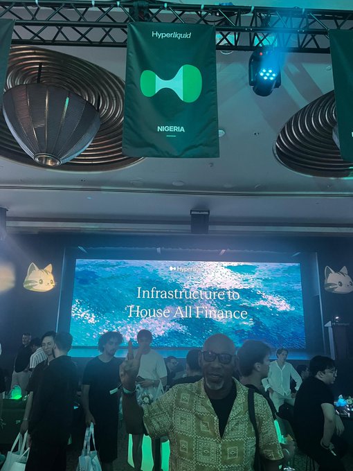 A man poses at a crypto event with a banner reading "Hyperliquid NIGERIA" hanging above and a screen behind him displaying "Hyperliquid - Infrastructure to House All Finance."