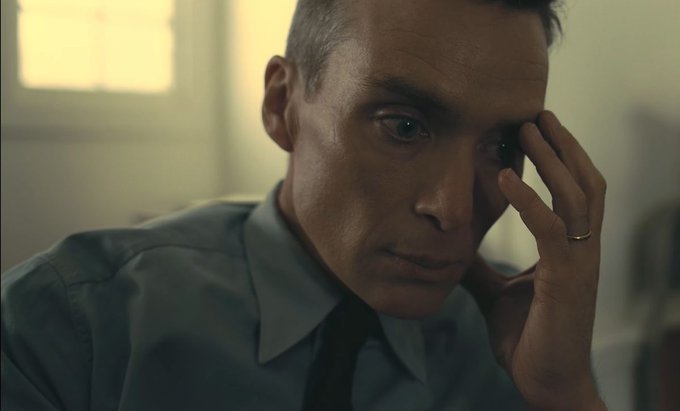 A cinematic close-up of a man in a light blue shirt and tie looking contemplative, with his hand resting against his face, wearing a ring.