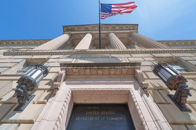 A low-angle view of the United States Department of Commerce building, featuring classical architecture, columns, ornate lamps, and an American flag waving against a blue sky.
