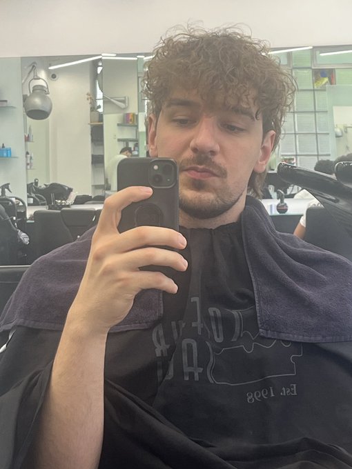 The image features a man sitting in a barber shop, taking a selfie with his cell phone. He is wearing a black shirt that says "Time for a new perm" and has curly hair. The photo captures him from behind, making him appear larger than life as he poses for the camera. The background of the image shows various barber tools and equipment, indicating that this is a professional setting like a barbershop.
