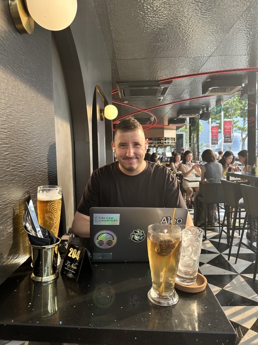 A man smiling at the camera while working on a laptop covered in stickers, seated at a cafe table with beer and iced drinks.