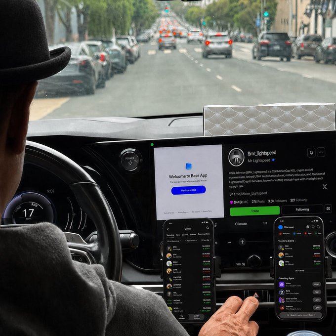 A view from inside a car showing the dashboard with a digital interface displaying a 'Welcome to Base App' prompt and what appears to be a social media feed, while the driver's hand reaches toward the screen on a city street.