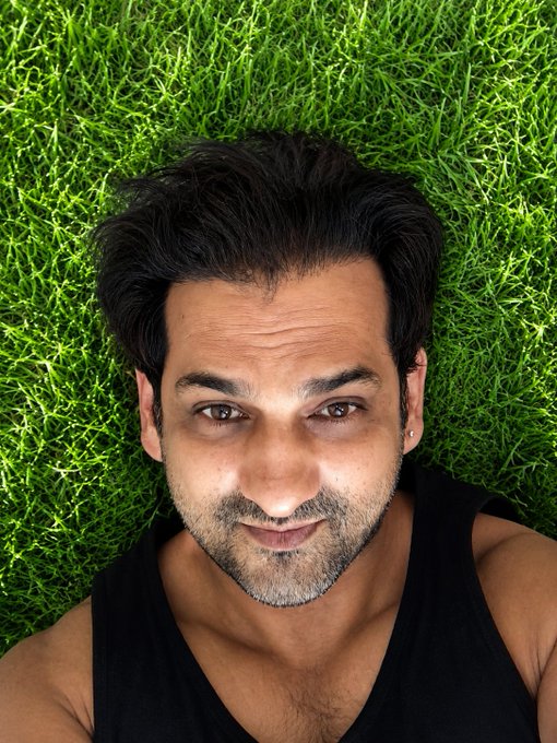 A close-up selfie-style portrait of a man with dark hair and a beard, lying on green grass and smiling at the camera.
