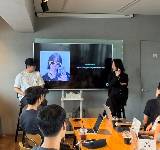 A woman with long black hair in a black outfit speaks at a small event, presenting a slide that displays her Twitter profile picture alongside text reading "guest speaker, joy building bitch @ founders.inc." The screen shows her profile wearing white sunglasses, while audience members with laptops watch in what appears to be a tech meetup at ZeroBase Korea.