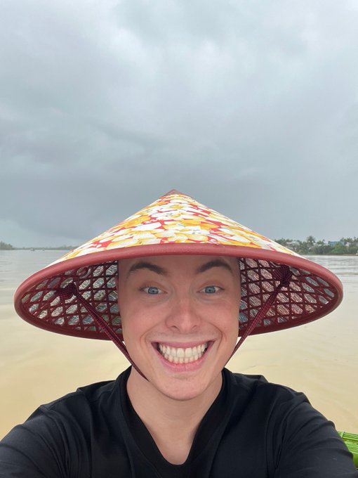 A smiling young woman wearing a colorful conical Vietnamese hat takes a selfie on a beach with cloudy gray skies and ocean visible in the background.
